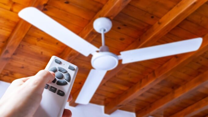 a hand points a remote control at a white ceiling fan hanging below a wood ceiling
