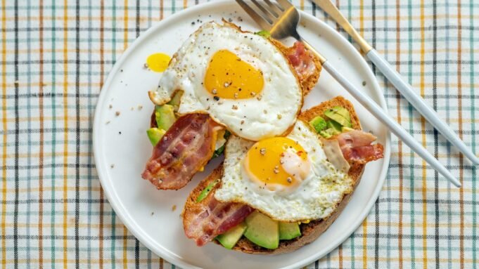 A fried egg, bacon, and avocado on top of toast on a white plate that sits on top of a checked tablecloth.